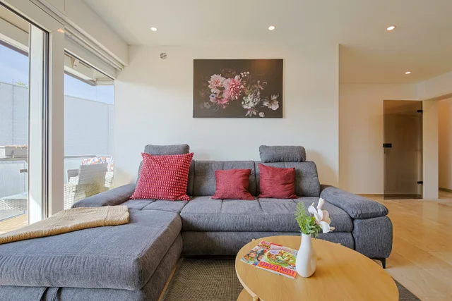 Wohnzimmer mit grauem Ecksofa, roten Kissen und Zugang zum Balkon.
